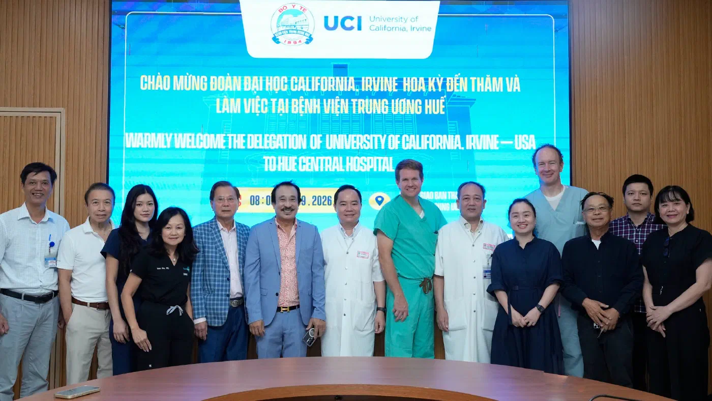 14 people from the UC Irvine delegation and Hue Central Hospital stand together in front of a large screen, one of them in black scrubs embroidered Trinh Nhu, M.D. The screen carries the hospital's emblem and the UCI, University of California, Irvine logo, and reads Chào mừng đoàn Đại học California, Irvine Hoa Kỳ đến thăm và làm việc tại Bệnh viện Trung ương Huế, then Warmly welcome the delegation of University of California, Irvine, USA to Hue Central Hospital.