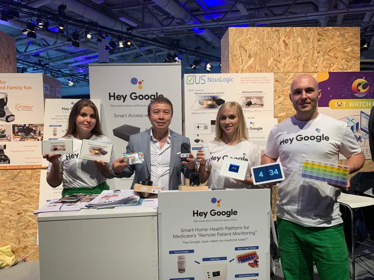Hoang Nhu at the Nouslogic stand at IFA Berlin with three Google brand ambassadors in Hey Google shirts, holding Nouslogic products: Watchman controller boxes, a smart medical card, a video doorbell, a pill bottle, a smart hub, a display clock and a seven day pill box. The booth signs read Hey Google, Smart Access, and Smart Home-Health Platform for Medicare's Remote Patient Monitoring, and the Nouslogic poster gives the stand number and the Berlin and Las Vegas dates.