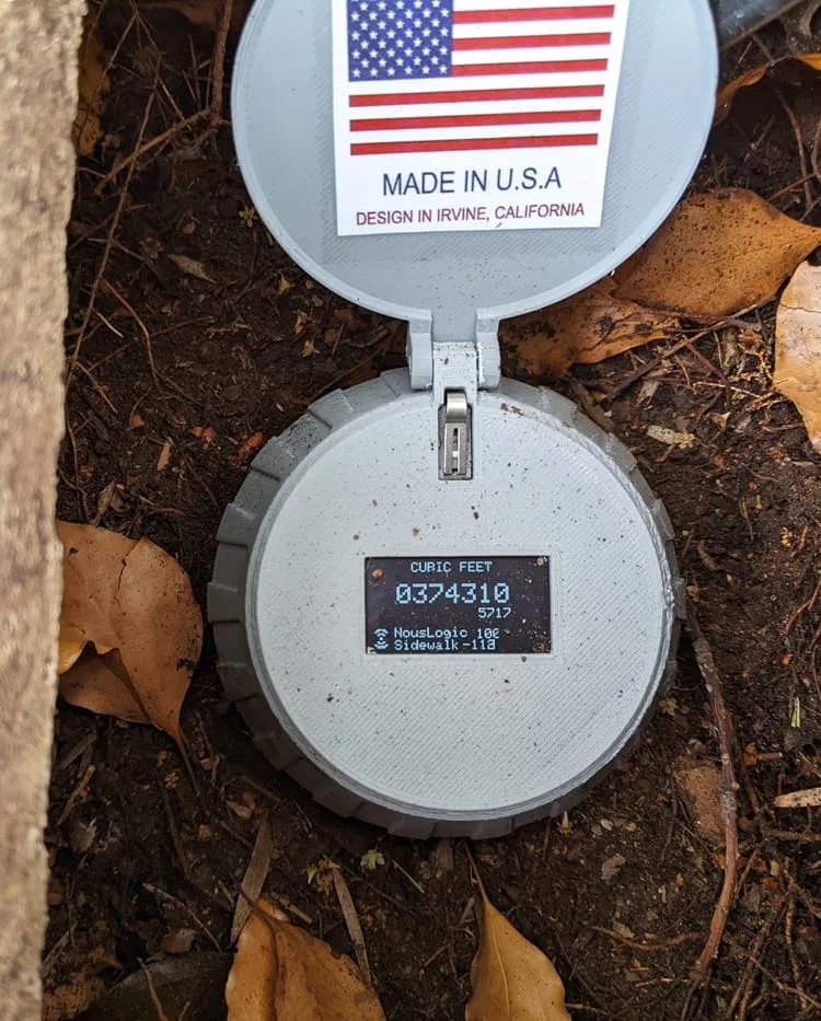 A Nouslogic smart meter reader in a ground box beside a path, its hinged lid standing open. The lid carries a US flag sticker printed MADE IN U.S.A and DESIGN IN IRVINE, CALIFORNIA. The unit's own display shows a reading in cubic feet, 0374310 above a smaller 5717, and under it a wireless symbol with NousLogic 102 and Sidewalk -118. Soil and dry leaves around it.