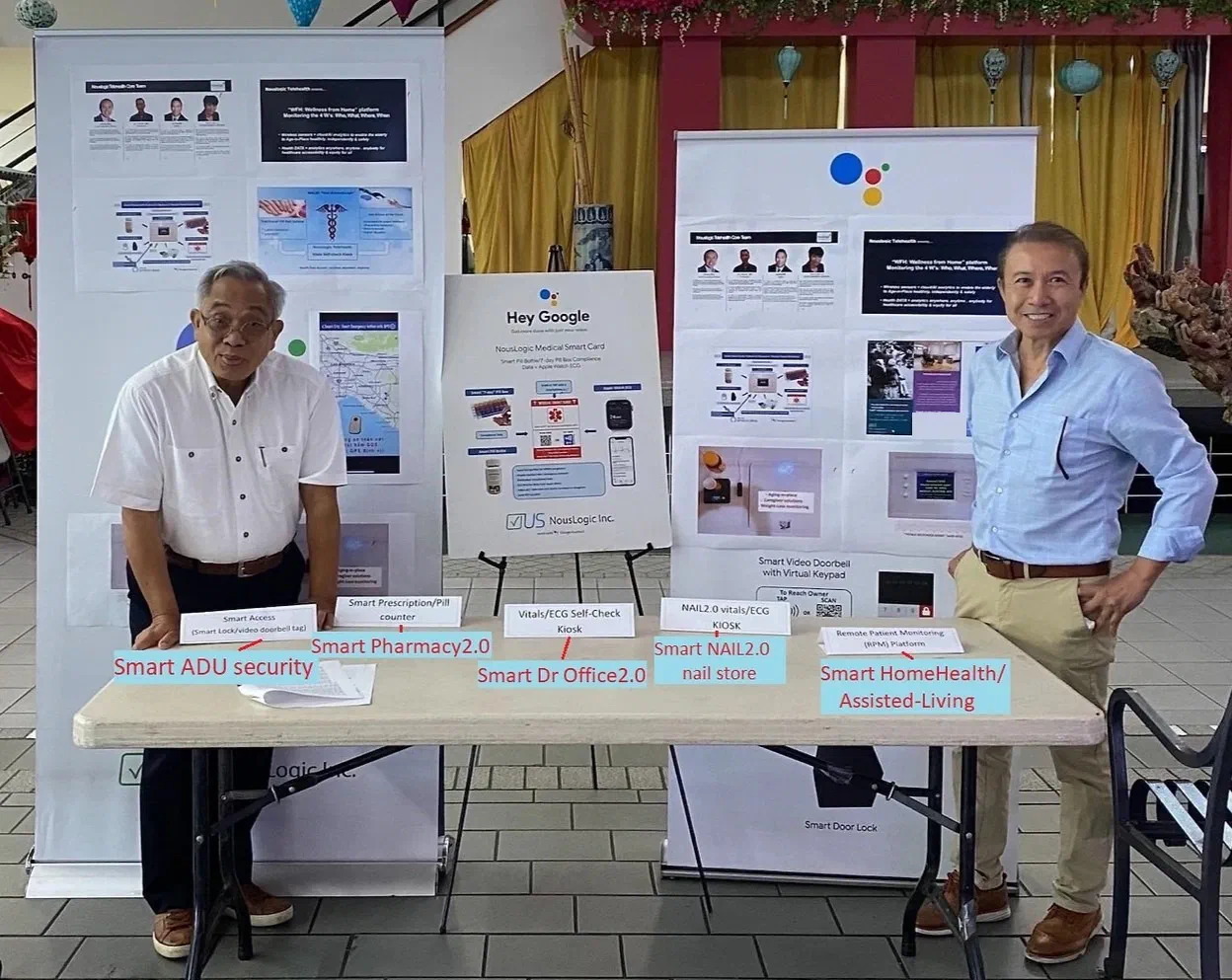 Hoang Nhu and the founder and developer of Asian Garden Mall behind a demonstration table inside the mall, in front of pull up banners for the Wellness From Home platform and a Hey Google easel board. Table cards read Smart Access, Smart Prescription and Pill Counter, Vitals and ECG Self-Check Kiosk, NAIL2.0 kiosk, and Remote Patient Monitoring Platform, with annotation arrows naming the Smart ADU security, pharmacy, doctor office, nail store and home health scenarios.