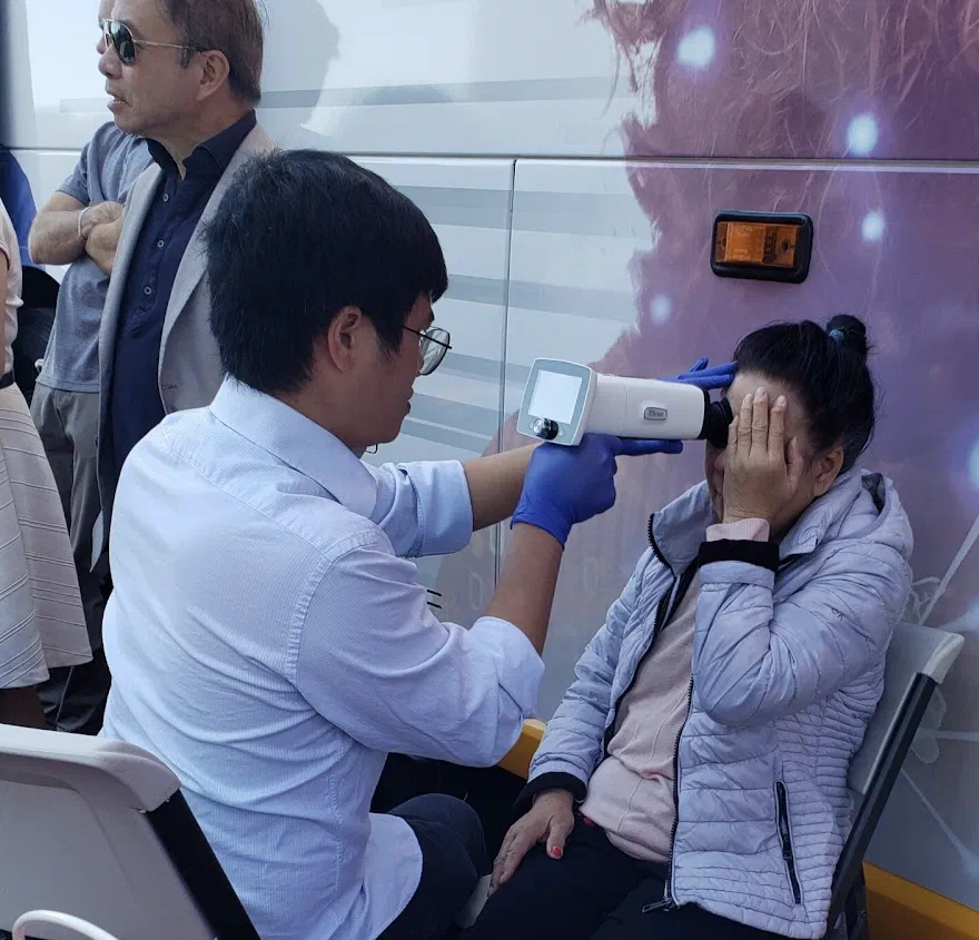 A technician in gloves holding a handheld retinal camera up to the eye of a woman seated beside the mobile clinic van, while she shields her other eye with her hand. Hoang Nhu stands watching at the left of the frame.