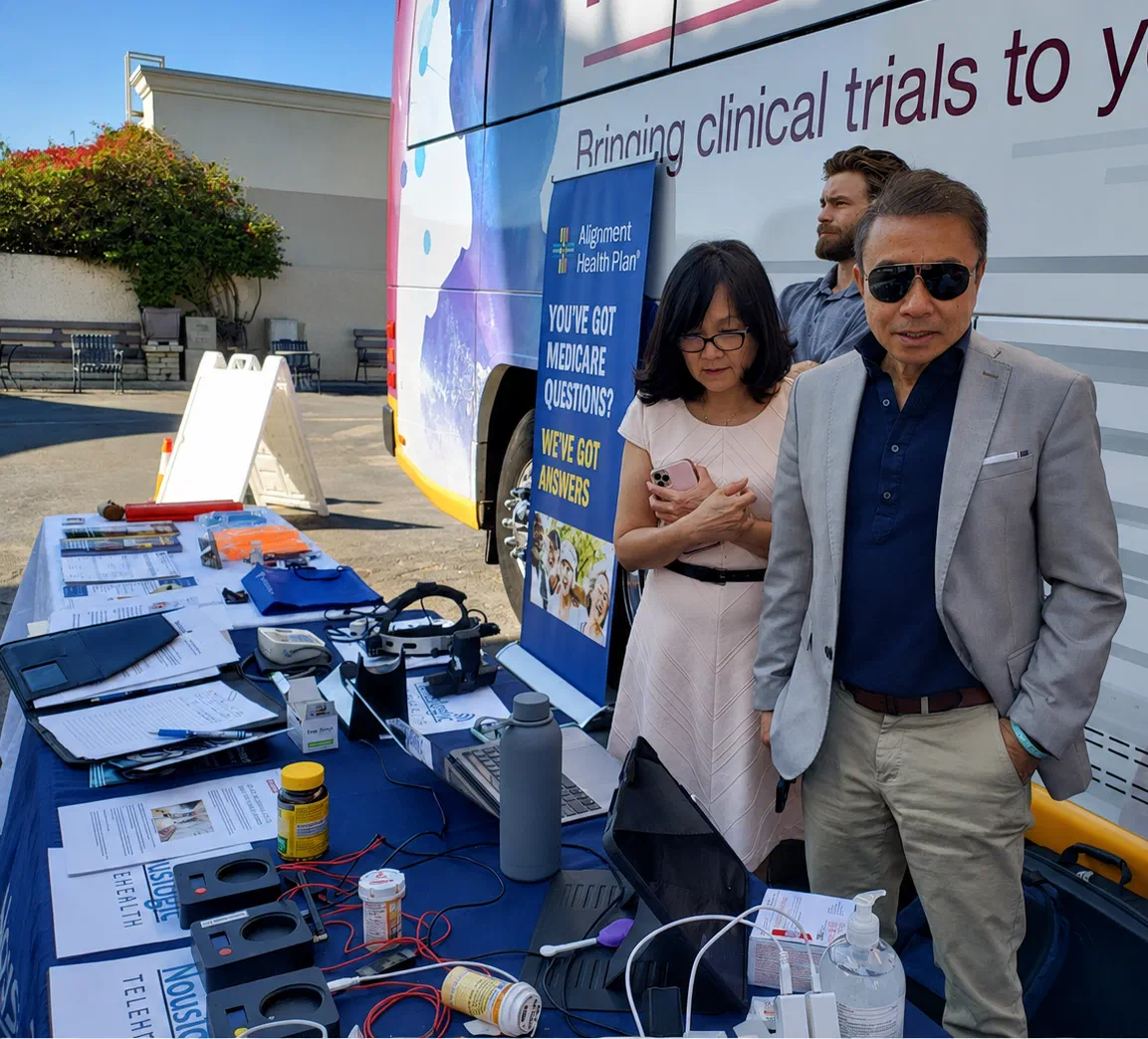 Hoang Nhu standing beside a mobile clinic van at an outdoor community health screening, next to a table laid out with blood pressure cuffs, monitoring devices and NousLogic Telehealth leaflets, with two colleagues standing behind him. The van reads Bringing clinical trials to you.