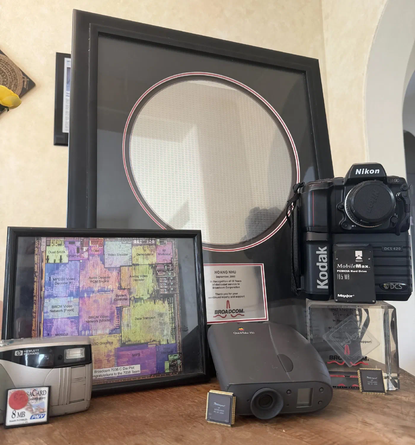A shelf of objects from 25 years of chip design: a framed 300mm silicon wafer; a framed Broadcom 7038 die plot with block labels such as MPEG2 Video Decoder, Audio Decoder PCM Engine and MIPS; a Broadcom plaque reading Hoang Nhu, September 2000, In Recognition of 10 Years of dedicated service to Broadcom Corporation; a Nikon N90 on a Kodak DCS 420 digital back with a 105MB MobileMax PCMCIA hard drive; an Apple QuickTake 150; an HP PhotoSmart camera with an 8MB flash card; a smaller Broadcom acrylic award; and 2 gold-pinned chip packages printed HP CASABLANKA 09 16 1994.