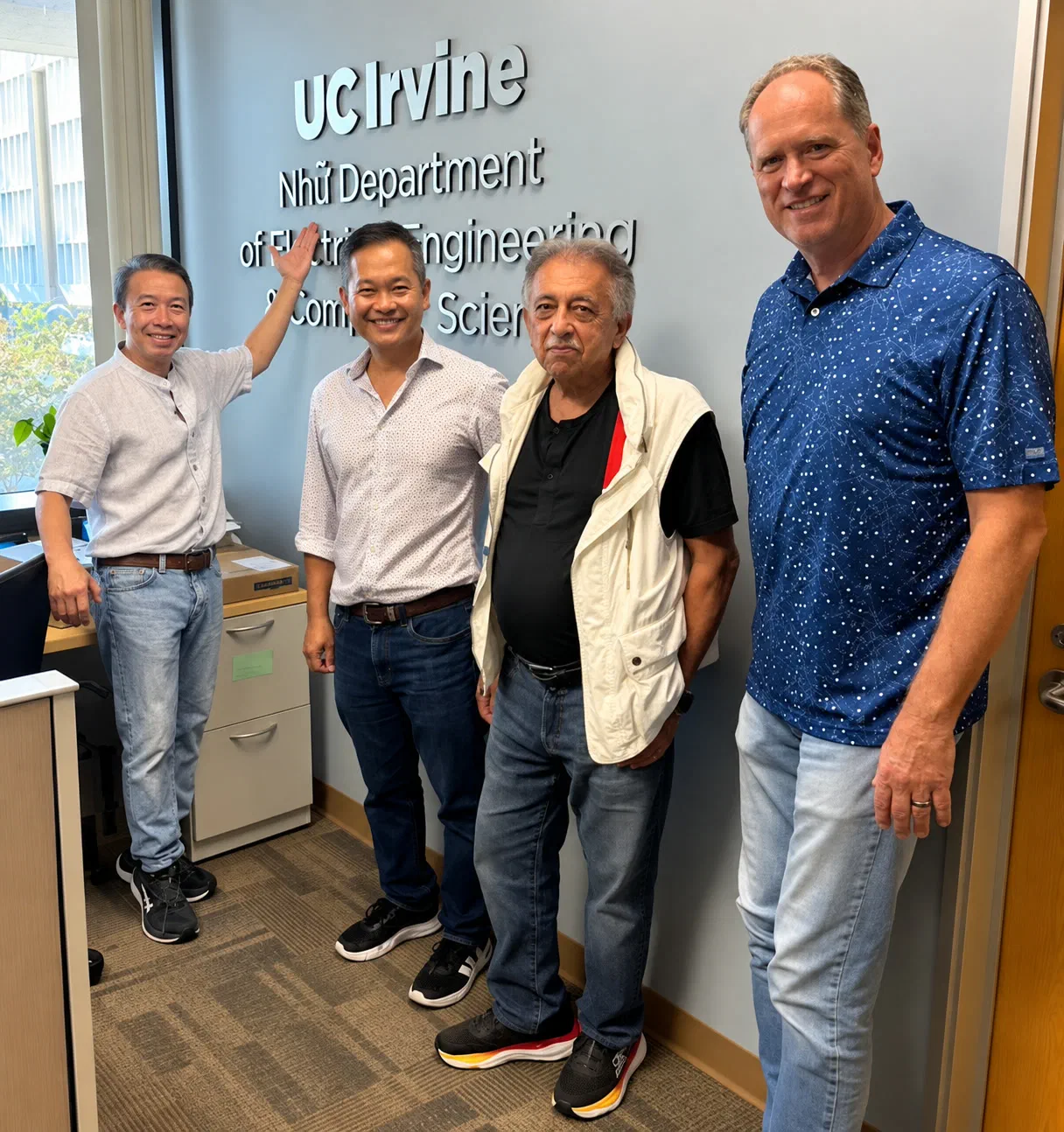 Four men stand in front of the department's name in raised white letters on a grey wall at UC Irvine, the one on the left reaching up to point at it. The lettering reads UC Irvine, Nhữ Department of Electrical Engineering & Computer Science, its lower two lines partly hidden behind them. A window on the left looks out onto trees.