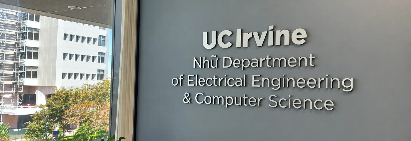The department's name in raised white letters on a grey wall indoors, reading UC Irvine, Nhữ Department of Electrical Engineering & Computer Science. A window on the left looks out onto a building under scaffolding and a row of trees.