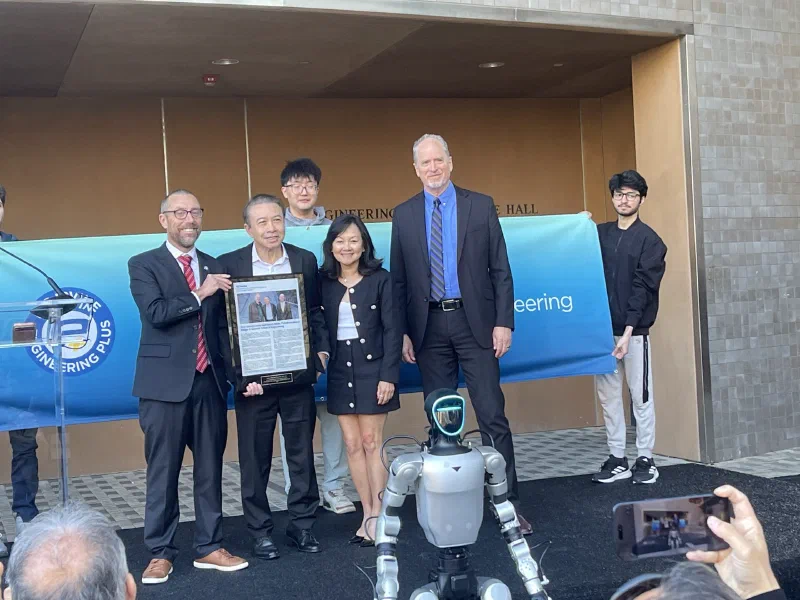 Hoang Nhu holding a framed print of the UC Irvine announcement, standing with three colleagues in front of a UC Irvine Engineering banner. A humanoid robot stands in front of the group and students hold the banner behind them.