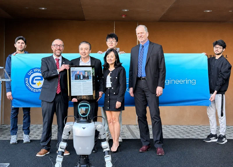 Hoang Nhu holding a framed print of the UC Irvine announcement, standing with three colleagues in front of a UC Irvine Engineering banner. A humanoid robot stands in front of the group and students hold the banner behind them.