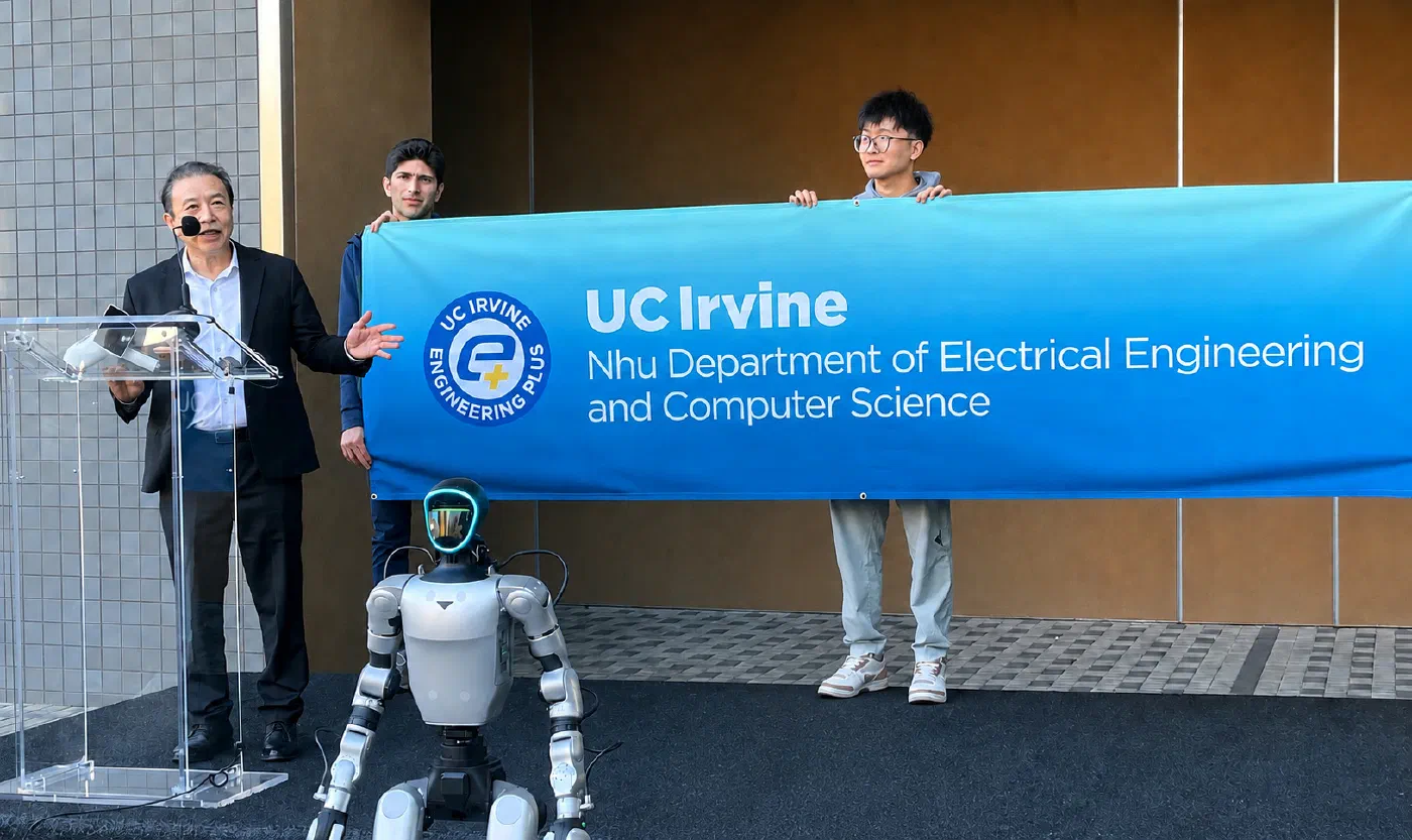 Hoang Nhu speaking at a clear lectern outdoors. Behind him students hold a large blue banner reading UC Irvine, Nhu Department of Electrical Engineering and Computer Science. A humanoid robot stands on the platform in front of the lectern.