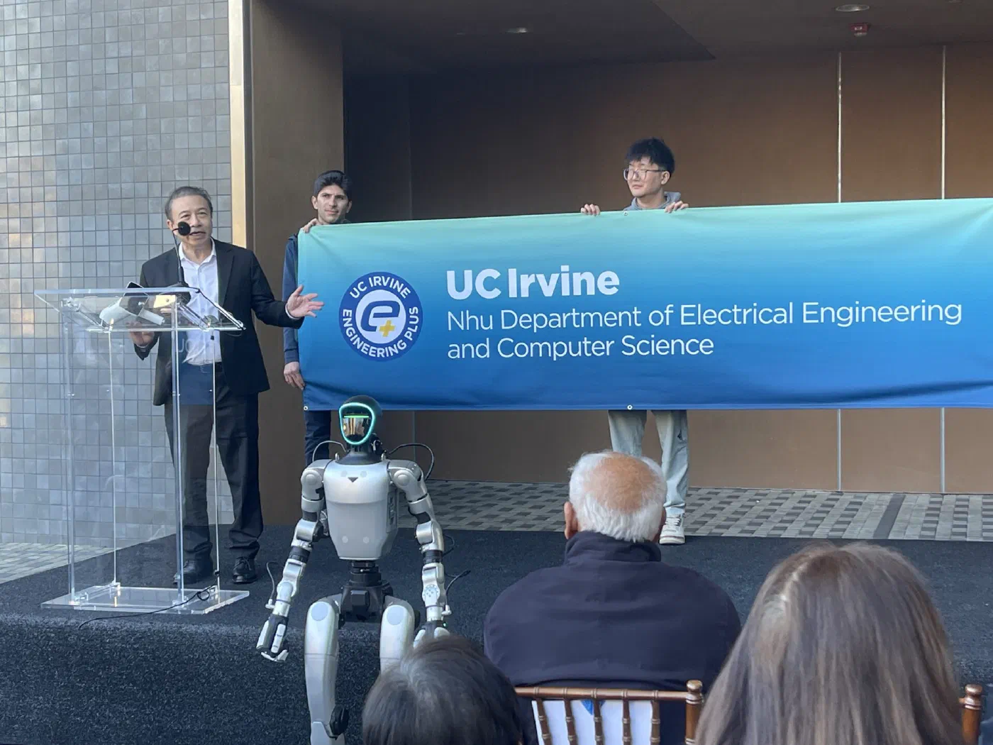 Hoang Nhu speaking at a clear lectern outdoors. Behind him students hold a large blue banner reading UC Irvine, Nhu Department of Electrical Engineering and Computer Science. A humanoid robot stands on the platform in front of the lectern.
